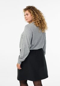 Frau mit lockigem Haar, die ein graues Langarmoberteil trägt, das in einen schwarzen Rock gesteckt ist, und die in einer dreiviertel Rückansicht vor einem weißen Hintergrund steht.