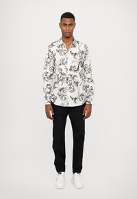 Camisa blanca de botones con un patrón floral negro en espiral, combinada con pantalones negros. Modelo usando zapatillas blancas, de pie frente a un fondo liso.