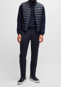 Laivaston sininen tikattu liivi korkealla kauluksella, yhdistettynä siniseen neulepuseroon ja tummiin housuihin, joita täydentävät mustat lenkkitossut valkoisilla pohjilla.