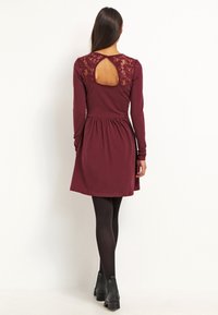Žena v burgundy dlouhých šatech s krajkou a slzovým výstřihem na horní části zad, s černými punčochami a černými kotníkovými botami, stojící.