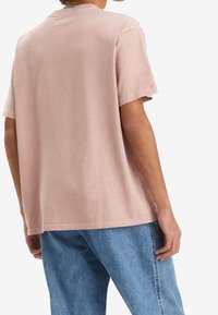 Světle růžové tričko s krátkým rukávem ve volném střihu, vyrobené z měkké tkaniny. Viditelné švy podél lemu a rukávů. Nosené s modrými džínami.