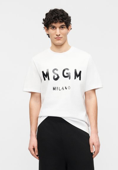 Jeune homme aux cheveux bouclés portant un t-shirt blanc MSGM Milano et un pantalon noir, debout devant un fond clair uni.
