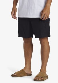 Black cargo shorts with side pockets, made of lightweight fabric, paired with brown slide sandals featuring a suede-like texture.