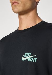 Sort bomulds t-shirt med et lysegrønt "Just Do It" logo og Nike swoosh på venstre brystområde. Klassisk crew neck design.