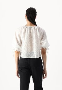 Bluză albă transparentă cu model în valuri, cu decolteu elastic și mâneci puff, asortată cu blugi negri. Textura pare ușoară și fluidă.