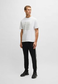 Hvid t-shirt med cirkulært grått grafisk design, parret med sorte joggers med hvide sidelister og sorte atletiske sko.