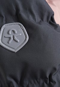 Črn material z teksturirano površino, ki vsebuje sivo logotip v obliki ščita z abstraktnim likom. Material izgleda gladko in rahlo sijoče.