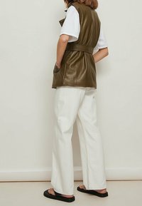 Oliwkowa kamizelka z ekoskóry z paskiem w talii, noszona na białej koszuli, zestawiona z białymi spodniami typu wide-leg i czarnymi klapkami.