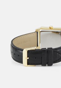 Montre rectangulaire dorée avec un bracelet en cuir noir à finition texturée et une boucle dorée. Les coutures visibles ajoutent des détails.
