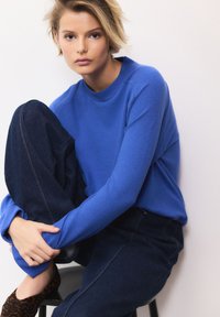 Maglione blu a maniche lunghe con scollo tondo abbinato a jeans in denim scuro, caratterizzato da una vestibilità morbida e dettagli di cuciture visibili.