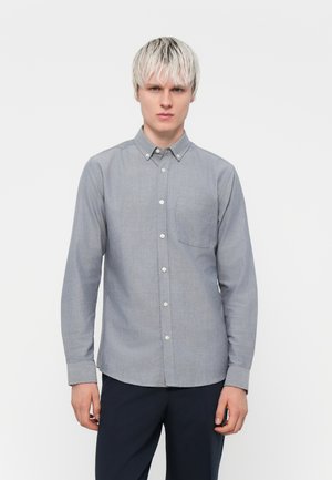 Camisa gris de botones con mangas largas, un bolsillo en el pecho izquierdo, cuello clásico y textura de tela ligera. Combinada con pantalones oscuros.