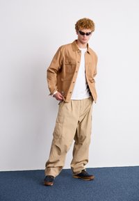 Beige bomull skjortejakke med knapper og brystlommer, sammen med beige cargo-bukser og svarte sko, mot en nøytral bakgrunn.