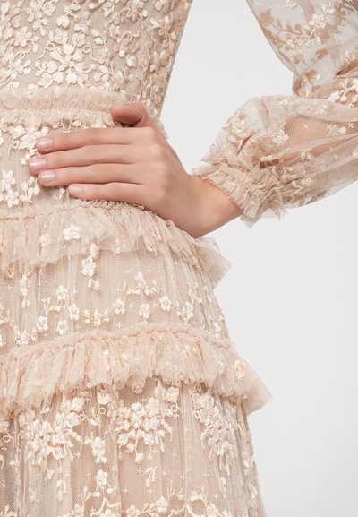 Kremo spalvos nėriniuotas suknelė su gėlių siuvinėjimu, sluoksniuotais volanais ties juosmeniu ir permatomomis pūstomis rankovėmis. Lengva, tekstūruota medžiaga.
