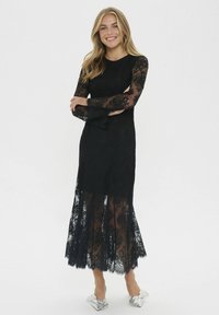 Juoda nėriniuota suknelė su ilgomis permatomomis rankovėmis ir sluoksniuotu nėriniuotu apačiu. Suknelė turi prigludusią krūtinės dalį ir permatomus sijonų sluoksnius, derinama su metaliniais bateliais.