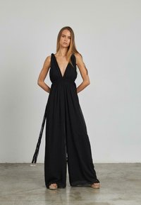 Mono negro con escote en V profundo, cintura fruncida y silueta de pierna ancha. Hecho de un tejido ligero con acentos de borlas laterales.