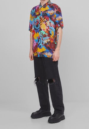 Persoon die een kleurrijk T-shirt met abstract patroon en korte mouwen draagt, zwarte gescheurde jeans en stevige zwarte schoenen, staand tegen een grijze achtergrond.