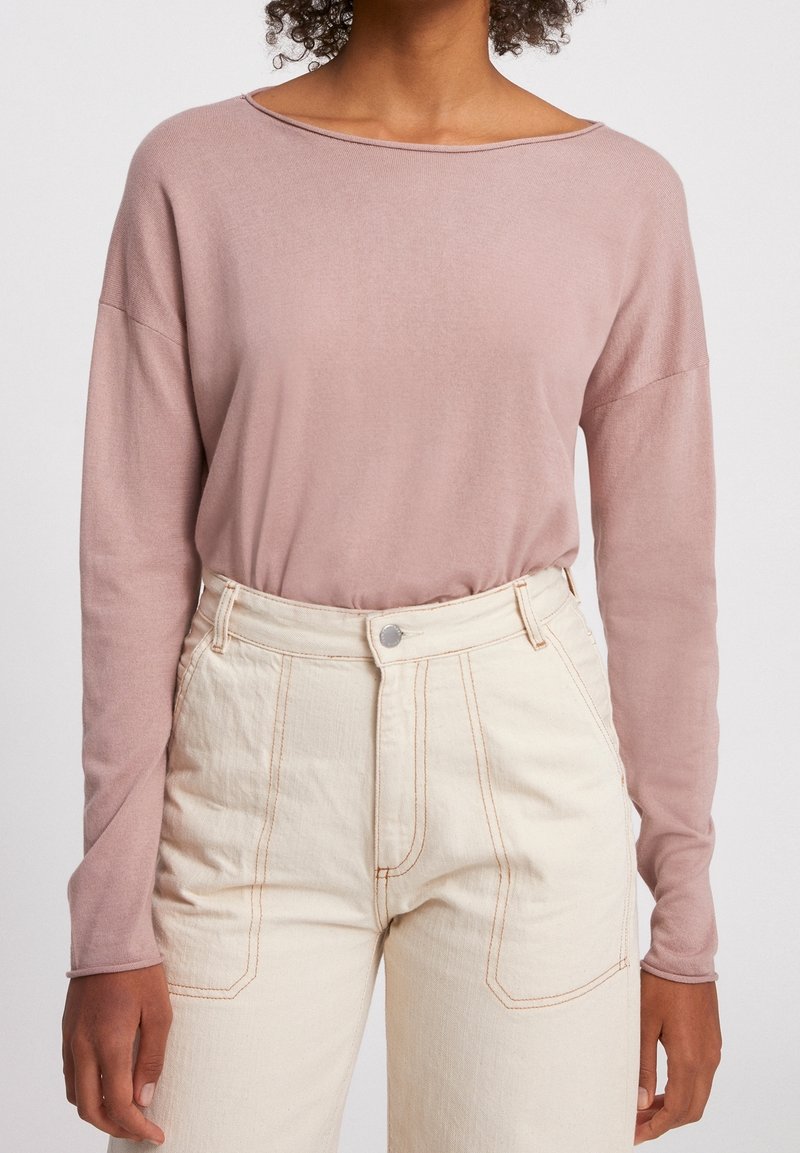 Personne portant un haut à manches longues mauve clair rentré dans un pantalon taille haute blanc cassé avec des coutures visibles et des poches avant.