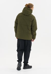 Pufferjack in olijfgroen, met een capuchon, gewatteerd ontwerp en elastische manchetten, gecombineerd met zwarte cargobroeken en zwarte sportschoenen.