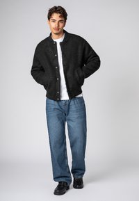 Black fuzzy bomber jacket with a ribbed collar, front snap buttons, and side pockets, paired with loose blue denim jeans and black shoes.