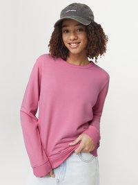 Roze sweatshirt met lange mouwen en geribbelde manchetten, gedragen met lichtblauwe denim. Een donkere pet met "LOVE MORE" in roze borduursel wordt ook gedragen.