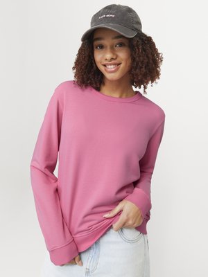 Roosa kapuutsiga sweatshirt pika varrukaga ja soonikkoes, selga pandud heledate siniste teksadega. Peas on tume müts, millel on roosa brodeeringuga kiri "LOVE MORE".