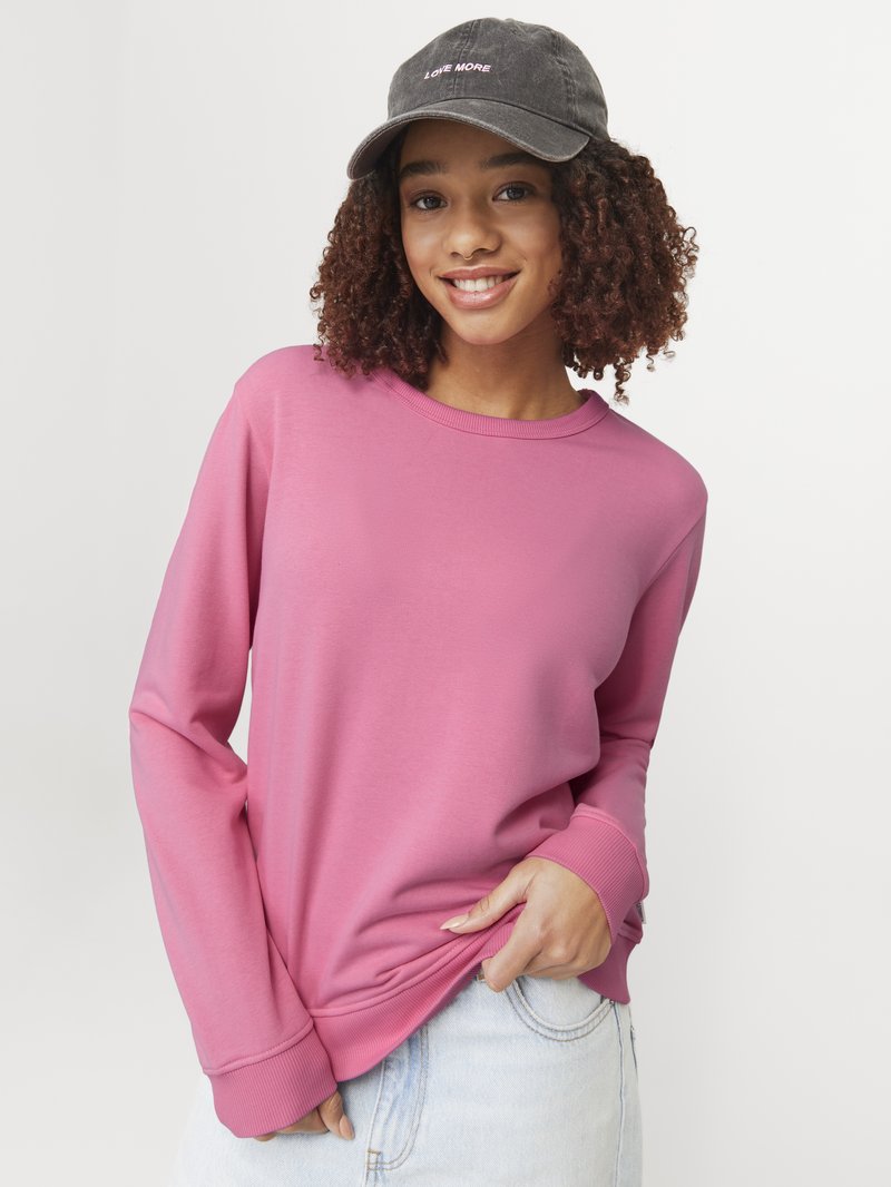 Roze sweatshirt met lange mouwen en geribbelde manchetten, gedragen met lichtblauwe denim. Een donkere pet met "LOVE MORE" in roze borduursel wordt ook gedragen.