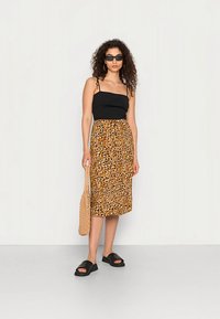 Top preto com alças finas, saia mostarda até ao joelho com estampa de leopardo, sandálias deslizantes pretas e uma mala bege trançada. Cabelo encaracolado e óculos de sol.