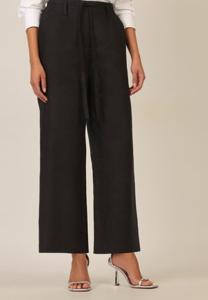 Frau trägt hoch taillierte, weit geschnittene schwarze Hose mit gebundener Taille, kombiniert mit silbernen Sandaletten mit Absatz und einer weißen Bluse.