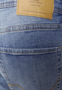 Jeans de denim azul claro com um patch de couro na cintura que exibe os detalhes da marca. Costuras visíveis e contornos dos bolsos.
