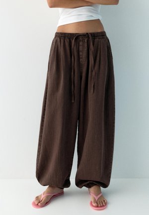 Persoană purtând pantaloni maro largi cu șnur și papuci roz, stând cu brațele încrucișate pe un fundal simplu.