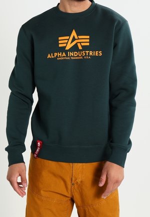 Person iført mørkegrøn Alpha Industries sweatshirt med orange logo og tekst, kombineret med sennepsgule bukser.