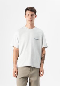 Joven con cabello castaño claro que lleva una camiseta gris y pantalones beige. Su brazo tiene un tatuaje y está de pie frente a un fondo blanco liso.