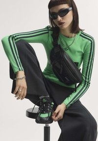 Young person with short brown hair wearing black sunglasses, green long-sleeve shirt with stripes, black pants, sneakers, and a black crossbody bag.