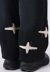 Pantalones negros con grandes detalles de cruces bordadas en beige, con bordes deshilachados. Combinados con zapatillas deportivas negras texturizadas.