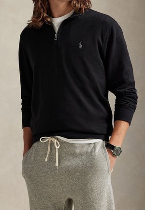Person trägt einen schwarzen Viertel-Zip-Sweatshirt mit kleinem Logo, hellgraue Jogginghose mit weißem Kordelzug und eine Armbanduhr am linken Handgelenk.