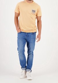 Lysegul t-shirt med en trykt blå lomme-logo, som er parret med blå jeans og hvide sneakers. Casual pasform, materiale i bomuld.
