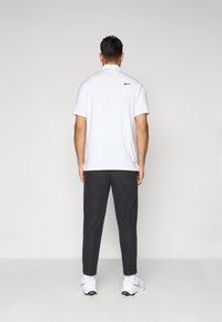 Mand, der står med ryggen til, iført en hvid Nike polo-shirt, sorte bukser, hvide Nike sneakers, på en ensfarvet grå baggrund.