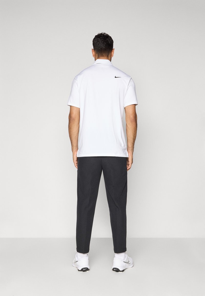 Man standing facing away, wearing a white Nike polo shirt, black pants, white Nike sneakers, on a plain gray background.