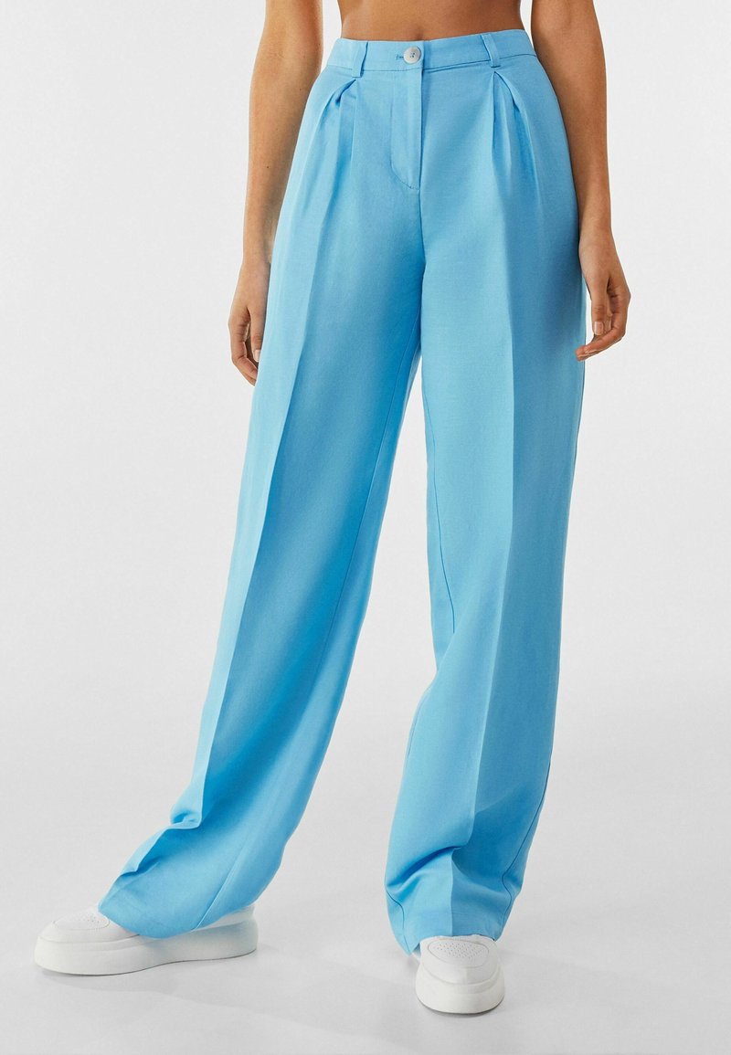 Light blue wide-leg trousers with pleats at the front, made of a smooth fabric. Features a button closure and pockets, paired with white sneakers.