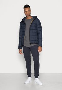 Marineblauwe pufferjack met capuchon, metallic rits en geribbelde manchetten; gecombineerd met een grijs T-shirt en donkere joggings; witte sneakers.