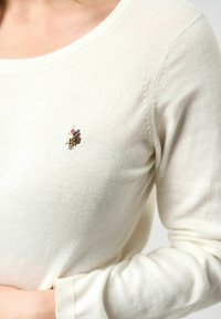 Pull crème clair à encolure ronde, texture fine en maille, avec un petit motif brodé sur la poitrine. Tissu doux avec des poignets côtelés.