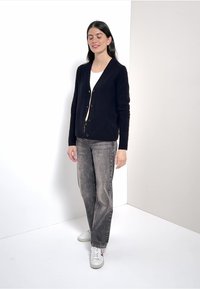 Marinblå cardigan med V-ringning och fem knappar, bärs över en vit topp; matchas med grå jeans och vita sneakers. En enkel bakgrund.