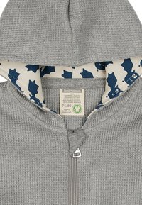 Grau Baumwoll-Hoodie mit einer strukturierten Außenschicht und blauer gemusterter Fütterung. Verfügt über einen Frontreißverschluss und ein Etikett, das die biologische Zertifizierung anzeigt.