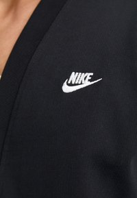Prenda de tela negra con texturizado en canalé que presenta un diseño de escote en V y un logo de Nike bordado en blanco en la parte superior izquierda.