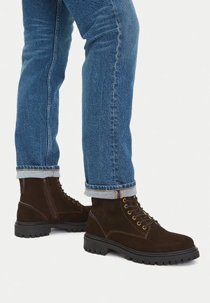 Legs wearing blue rolled-up jeans and dark brown suede lace-up boots with thick black soles on a white background.
