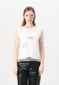 Maglietta bianca con maniche trasparenti, caratterizzata dal testo "Rebel" e appunti scritti a mano. Il fondo ha un orlo trasparente. Pantaloni in finta pelle nera.