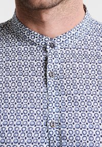 Skjorta med knäppning framtill och bandkrage, tillverkad av tyg med vit botten och marinblå cirkulära mönster, avslutad med mörka knappar.