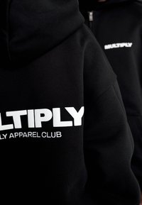 Sorte hættetrøjer lavet af blødt stof med fed hvid tekst "MULTIPLY" og "APPAREL CLUB" på forsiden, med en afslappet pasform og snore til hætten.