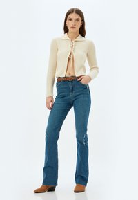 Ribbed cream cardigan with collar and tie closure, paired with blue flared jeans featuring distressed accents. Brown belt and suede boots.