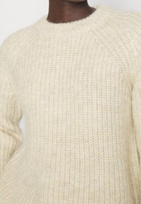 Dunkelhäutige Person trägt einen dicken, cremefarbenen, gerippten Strickpullover mit Rundhalsausschnitt.